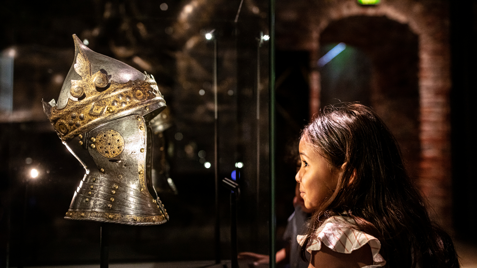 Två barn som står vid en monter i museet och tittar på en rustningshjälm