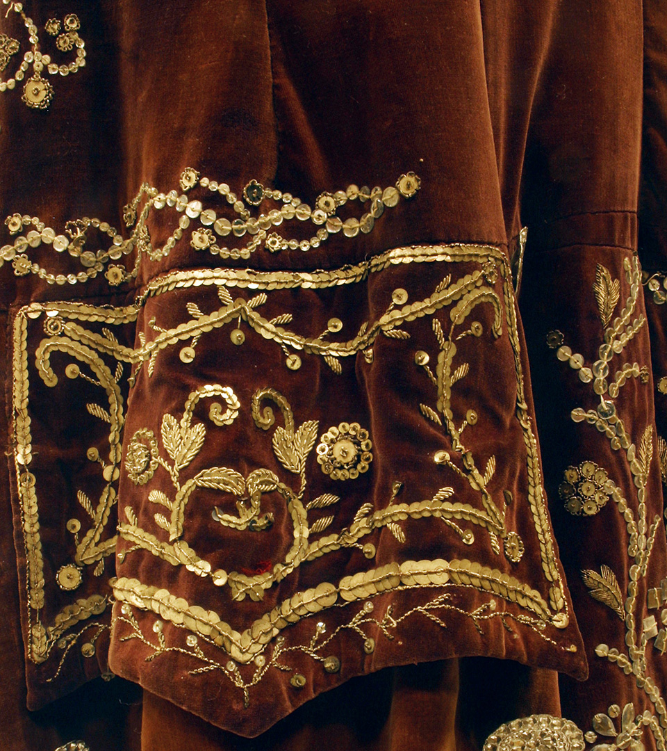 Detaljbild av en mansrock av auberginefärgad sammet med paljettbroderier i silver.