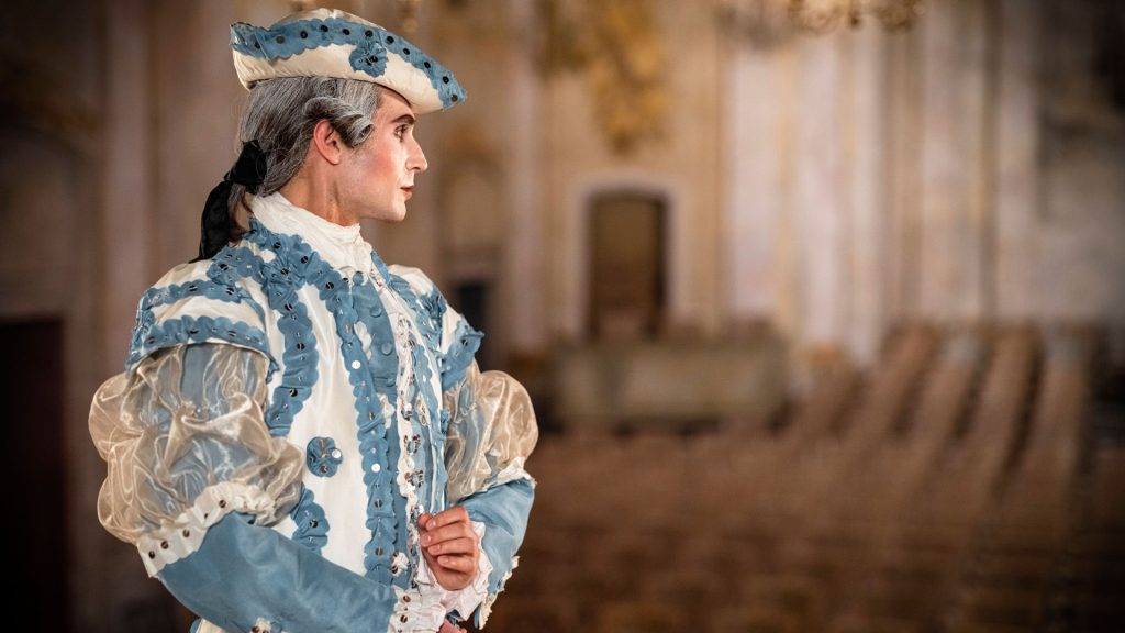 En person historiskt sminkad, med uppsatt peruk och klädd i en replika av Gustav III:s blåa dräkt. Personen står på en scen och blickar ut mot publiken.