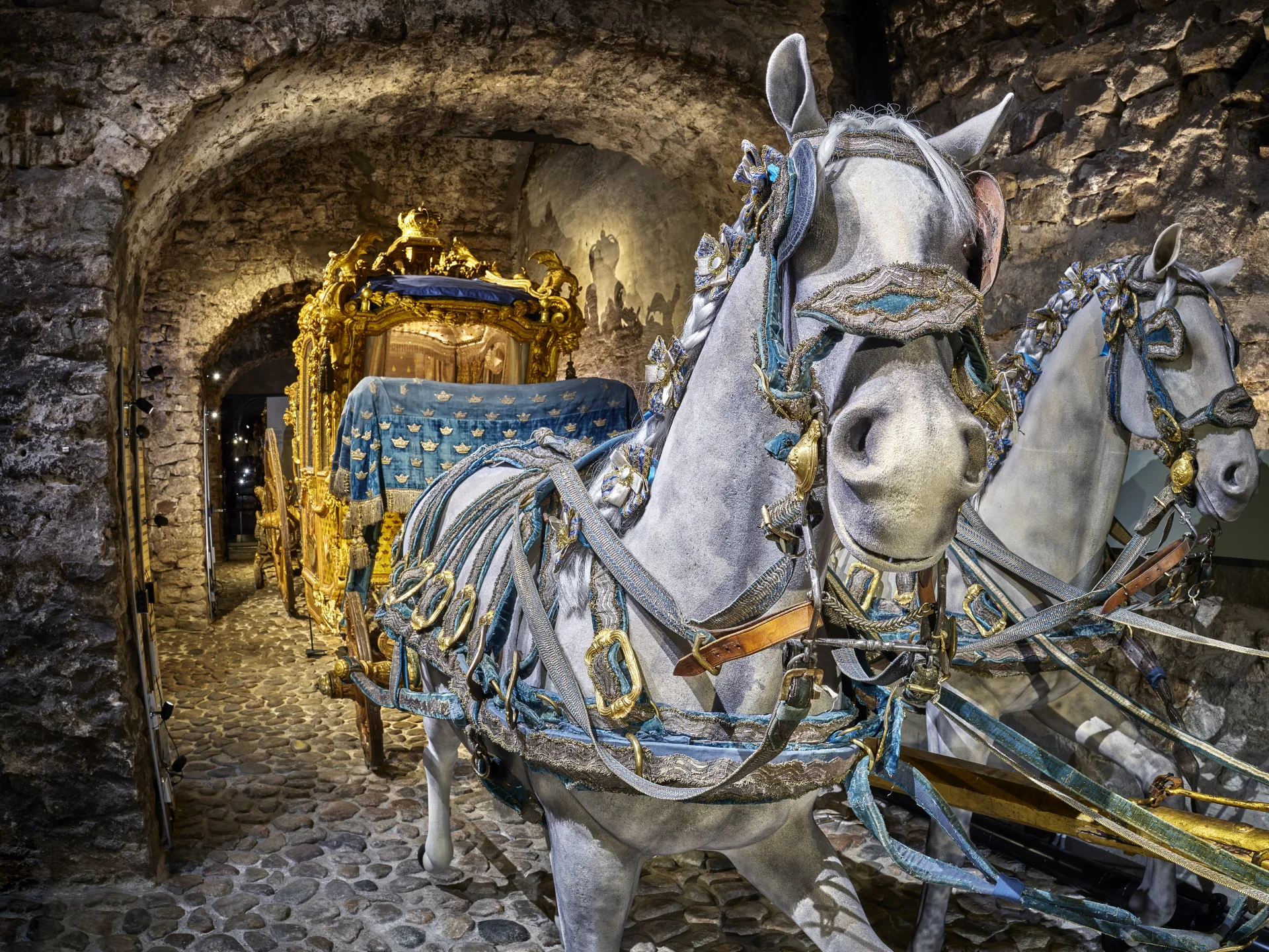 A lavish royal horse-drawn carriage in a stone-vaulted room with a cobblestone floor, pulled by two white-painted horse statues adorned with blue and gold harnesses. The carriage is richly decorated in gold and covered with a blue fabric embroidered with golden crowns. The photo is taken indoors in a historical exhibition setting, likely a museum.